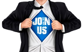 Man tearing open his suit jacket and shirt, revealing a blue t-shirt with white letters saying, "Join Us"
